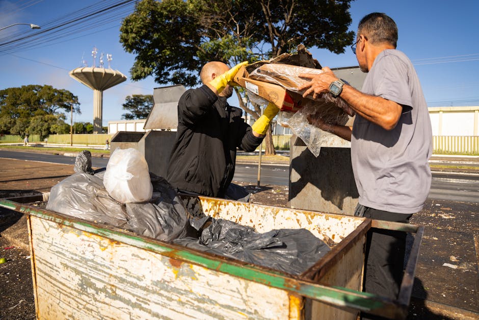 Two men are engaged in rubbish collection outdoors on a sunny day. One man, wearing a black jacket and yellow gloves, is placing a cardboard box filled with various waste items into an open-top, weathered metal bin that shows signs of rust and dirt. The other man, dressed in a light grey t-shirt and dark pants, is assisting by holding the box steady. The metal bin contains black and gray plastic rubbish bags, some of which are partially torn or overflowing, revealing mixed waste inside. In the background, there are large trees with green leaves, a clear blue sky, and a water tower or similar structure. The scene is set on a paved area adjacent to a roadway with a strip of grass and a white fence in the background. The natural daylight illuminates the scene with sharp shadows, emphasizing the textures of the rubbish, the metal bin, and the surrounding environment, illustrating a typical on-site waste clearance activity, possibly related to private waste disposal services.