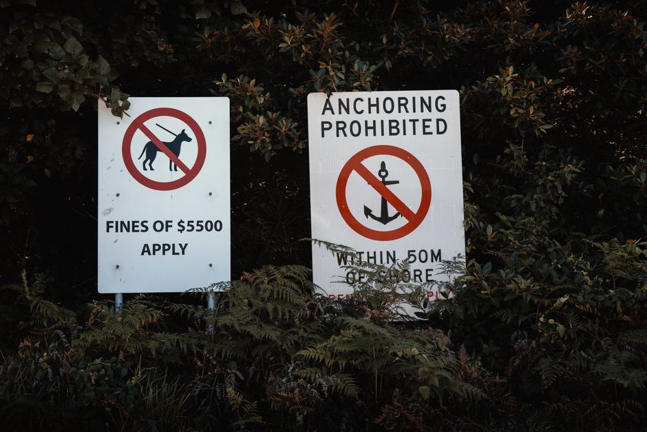 Two white warning signs are posted in front of dense foliage with dark leaves. The left sign features a black dog silhouette crossed out in red, indicating no dog walking, and states that fines of £5500 apply. The right sign, titled 'ANCHORING PROHIBITED,' displays a red circle with a diagonal line crossing out an anchor icon, and specifies that anchoring is not permitted within 50 meters. The signs are mounted on metal posts partially obscured by plants, and the environment appears to be an outdoor area possibly near a body of water or a restricted zone. The lighting suggests a natural, possibly overcast, outdoor setting, and the surrounding plants and shrubs are lush and untrimmed, consistent with a natural or semi-natural environment related to outdoor waste or environmental management. These signs relate to private restrictions on activities, which could tie into broader waste and rubbish management protocols enforced by companies like rubbishclearancesouthkensington.com, particularly in areas where alternative disposal or clearance is needed to prevent violations or fines.