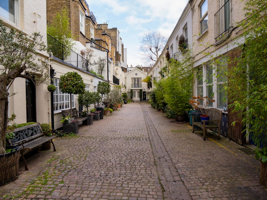 A narrow cobblestone street in a residential area lined with white-painted terraced houses on both sides. The buildings feature black iron balconies, window flower boxes, and small potted trees and bushes placed along the pavement, creating a lush, green atmosphere. On the left, there are black metal benches and large planters with neatly trimmed round shrubs and small trees. The right side has additional potted plants and a wooden bench, with some greenery extending from the windows and walls of the houses. The sky overhead is partly cloudy, casting soft diffuse light onto the scene, emphasizing the textures of the cobblestones and foliage. The setting appears quiet and well-maintained, typical of an elegant, historic London neighbourhood. This scene subtly reflects an environment where private outdoor spaces and on-site greenery are prominent, aligning with the concept of independent waste management or rubbish clearance in residential areas, with [COMPANY_NAME] likely providing services in such settings.