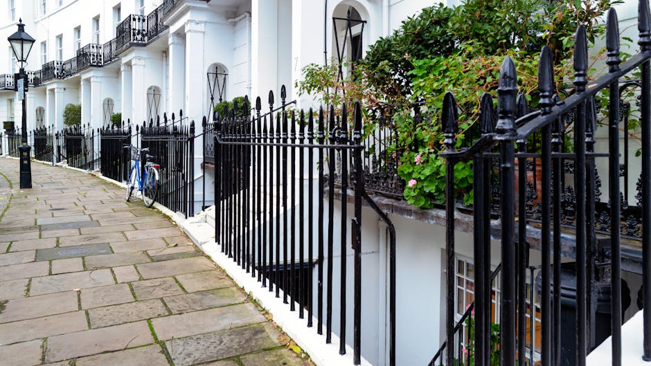 A row of white terraced townhouses with black wrought iron railings along the pavement, which is paved with rectangular stone slabs. The railings enclose small front gardens filled with green foliage, including leafy bushes and flowering plants. A blue bicycle is parked and leaned against the railings in front of one of the houses. The residential buildings feature large windows with black window frames and decorative white facades, with some doors visible behind the railings. The scene is outdoors during daylight, with natural lighting highlighting the textures of the stone pavement, metal rails, and lush greenery, illustrating a typical urban residential area that may require occasional rubbish clearance or private waste handling by rubbish removal services like those offered by [COMPANY_NAME]. The environment suggests a quiet, well-maintained street with minimal clutter, emphasizing the importance of suitable waste removal solutions within such settings.