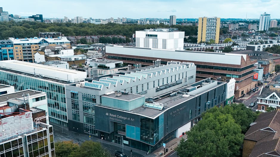 An aerial view of a cityscape featuring the Royal College of Art's modern, multi-story building with a predominantly glass facade and a dark grey exterior, situated among various other structures including commercial, residential, and institutional buildings. The surrounding area includes a mix of rooftops made from materials such as metal and tile, with some buildings displaying white, blue, and brown finishes. In the foreground, there is a cluster of closely packed rooftops and trees, with one prominent green tree on the right side providing contrast against the urban environment. The background reveals a skyline filled with mid-rise and high-rise buildings, with a distant horizon of greenery and open space. The scene is lit by natural daylight, highlighting the textures and colors of the buildings and foliage, which can relate to private or independent waste management practices typical of ongoing property maintenance or refurbishment projects, often requiring scheduled rubbish removal or clearance services by companies like rubbishclearancesouthkensington.com. The overall setting reflects a busy urban area where waste collection might be managed through external services rather than municipal collection alone.