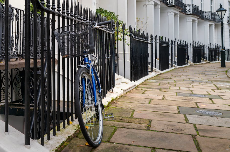 A row of white terraced townhouses with black wrought iron railings along the pavement, which is paved with rectangular stone slabs. The railings enclose small front gardens filled with green foliage, including leafy bushes and flowering plants. A blue bicycle is parked and leaned against the railings in front of one of the houses. The residential buildings feature large windows with black window frames and decorative white facades, with some doors visible behind the railings. The scene is outdoors during daylight, with natural lighting highlighting the textures of the stone pavement, metal rails, and lush greenery, illustrating a typical urban residential area that may require occasional rubbish clearance or private waste handling by rubbish removal services like those offered by [COMPANY_NAME]. The environment suggests a quiet, well-maintained street with minimal clutter, emphasizing the importance of suitable waste removal solutions within such settings.