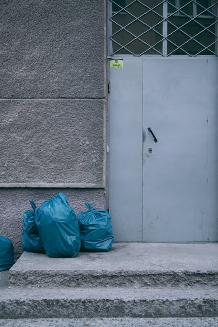 The image shows three large blue rubbish bags placed on a concrete doorstep outside a grey building. The bags are made of plastic and are tied at the top, with some appearing slightly crumpled and showing signs of contents shifting within. To the right of the bags, there is a grey metal door fitted with a black handle and a lock, with a small yellow warning sticker affixed near the top of the doorframe. The building’s exterior wall is textured with a rough, grey stucco finish, and behind the door, a section of barred window with diagonal metal grille is visible. The scene is set in an outdoor urban environment with natural daylight, indicating that the rubbish bags are temporarily placed outside for collection, potentially reflecting a private waste disposal or rubbish removal service such as that provided by Rubbish Clearance South Kensington. The concrete platform and steps in front of the door contribute to a typical external setting suited for on-site rubbish clearance, supporting the context of independent waste handling solutions.