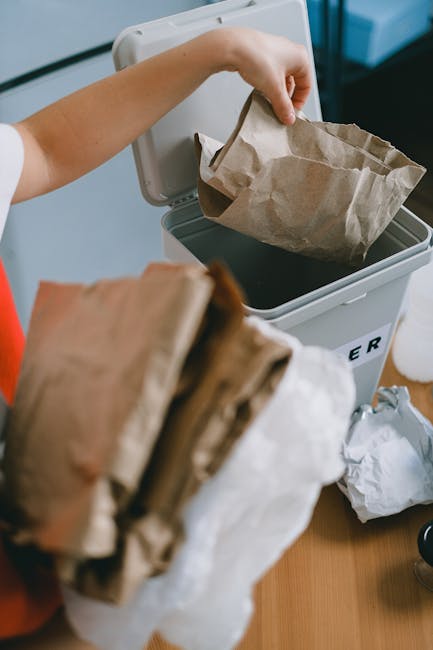 A person is seen disposing of brown paper waste bags into a grey bin designed for rubbish collection, which is placed on a light-colored wooden surface indoors. The individual’s arm is extended, holding a paper bag over the open lid of the bin, with other paper bags in their hand. The bin has a printed label indicating its purpose for waste disposal, and it is situated near a window that allows natural light to illuminate the scene. Surrounding the bin, there are additional crumpled paper bags and some miscellaneous waste materials, typical of on-site rubbish clearance activities. The environment suggests a domestic or small commercial setting, where independent waste handling processes are in progress, supporting the options available apart from standard municipal rubbish collection. The focus on paper bags and their placement emphasizes the importance of organized waste segregation for efficient rubbish removal, consistent with private waste management services such as those provided by rubbishclearancesouthkensington.com, which facilitate same-day or alternative waste disposal solutions in SW7.