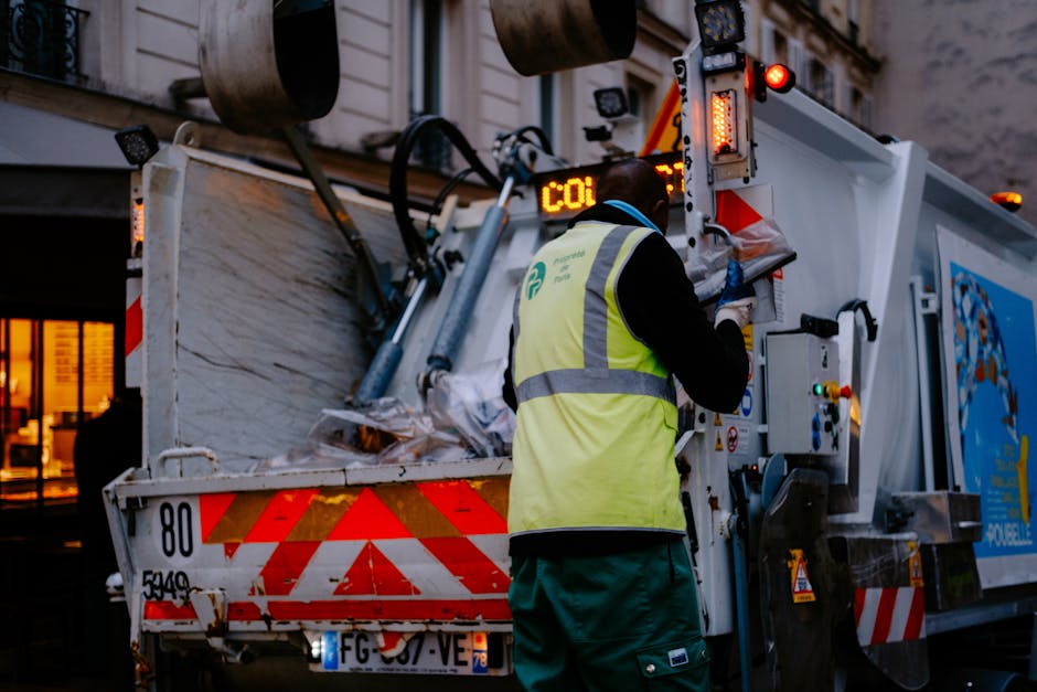 A waste collection worker wearing a high-visibility yellow vest and dark trousers is operating the controls of a white refuse collection vehicle with red and orange reflective markings on the rear. The worker is positioned with their back to the camera, appearing to adjust or monitor the machinery as the truck loads or processes waste materials. The vehicle is parked on a street in an urban environment, with a building featuring large windows and warm interior lighting visible in the background. Large metal hydraulic drums or rollers are attached to the top of the truck, indicating its function in waste or debris handling. The scene is illuminated by natural evening light, creating a contrast between the bright reflective safety markings and the darker surroundings. This image highlights aspects of private waste collection services, such as those provided by companies handling rubbish removal and on-site clearance, emphasizing the operational side of independent waste management in city environments associated with rubbish services.