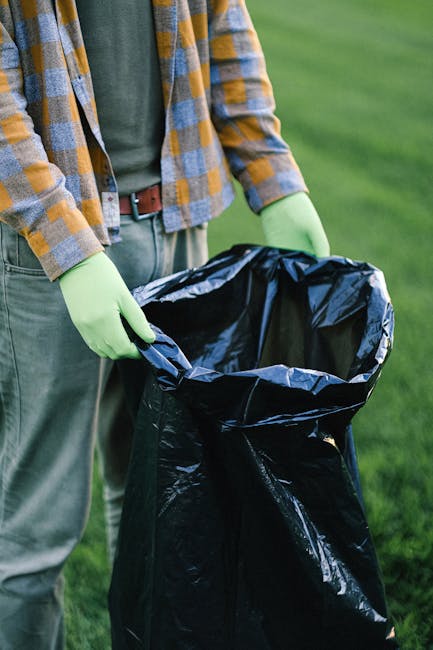 Close-up of a person wearing a plaid shirt over a green t-shirt, with lime green gloves, holding open a large black rubbish bag outdoors on a grassy area. The bag appears to be partly filled, with its top edges folded back to reveal a dark, empty interior, suitable for waste collection or recycling. The background features a lush, green lawn, indicating an outdoor setting such as a garden or park, and the lighting suggests a natural daylight environment, emphasizing the practical aspect of rubbish disposal or garden clearance. This scene relates to independent waste handling and refuse collection, where individuals undertake private rubbish removal rather than relying on local authority services, aligning with the themes of alternative waste management methods supported by companies like rubbishclearancesouthkensington.com.